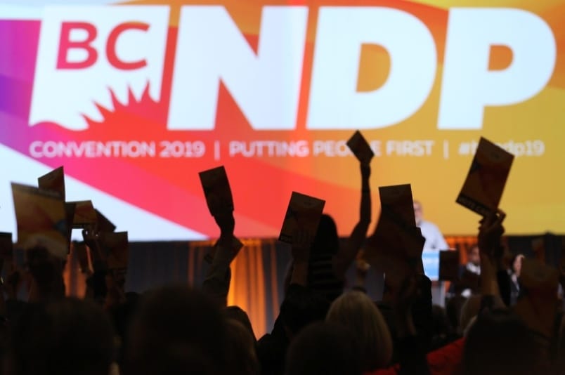 hands holding up voting cards in front of a large overhead screen displaying NDP logo